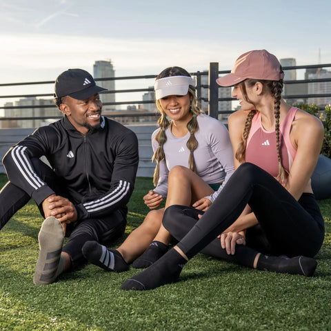 Three people sitting on grass wearing athletic wear and caps, smiling and conversing outdoors on a rooftop.