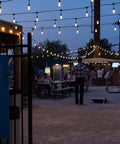 Cozy Cafe Style Outdoor String Lights With 48 Ft Black Cord 24 Shatterproof Acrylic Edison Bulbs Weatherproof For Patio Deck Garden Bars And Cafes Warm White Ambience For Indoor Or Outdoor Parties in outdoor evening setting with people gathered under lights.