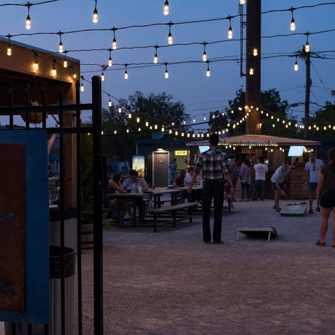 Cozy Cafe Style Outdoor String Lights With 48 Ft Black Cord 24 Shatterproof Acrylic Edison Bulbs Weatherproof For Patio Deck Garden Bars And Cafes Warm White Ambience For Indoor Or Outdoor Parties in outdoor evening setting with people gathered under lights.