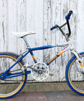 Vintage BMX bike with blue and white frame, blue tires, and swallow shaped handlebar set against wooden background.