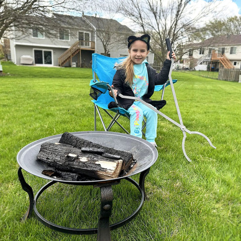 Child holding 40 inch stainless steel fire tongs sitting by fire pit on grass, ready for campfire use.