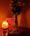 Dimmable Pink Himalayan Salt Lamp With Handcrafted Wooden Base next to a vase of flowers on a dimly lit table.