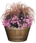 Whiskey Barrel Planter With Distressed Oak Finish And Antique Pewter Bands Yard Ready Durable UV Coated Resin Outdoor Drainage Holes Large Top Diameter 20.5 Inches Height 12.5 Inches Bottom Diameter 14 Inches filled with pink flowers and ornamental grass.