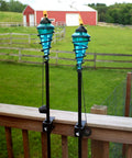 Two Pack Glass Patio Torch Set, tall outdoor torches on wooden deck with blue glass vessels and metal casings in garden setting.
