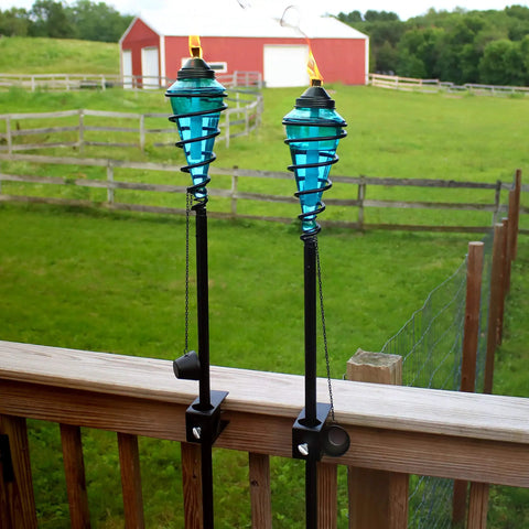 Two Pack Glass Patio Torch Set, tall outdoor torches on wooden deck with blue glass vessels and metal casings in garden setting.