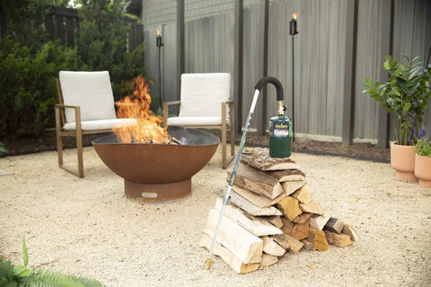 Self Igniting Outdoor Torch, 20,000 BTU High Output Flame, 36 Inch Handle leaning on wood near a fire pit