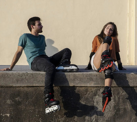 Two people wearing roller blades sitting on a ledge, enjoying a sunny day in casual attire with knee pads.