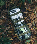 Rhino Rescue survival first aid kit in green MOLLE pouch opened on forest floor, showing bandages, tape, and tools.