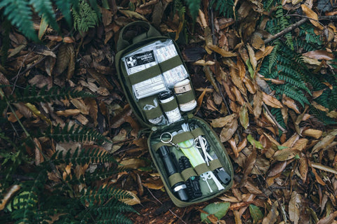 Rhino Rescue survival first aid kit in green MOLLE pouch opened on forest floor, showing bandages, tape, and tools.