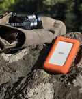 Rugged USB-C Portable External Hard Drive 5TB for Mac and PC, Drop Resistant, placed outdoors on a rock near a camera bag