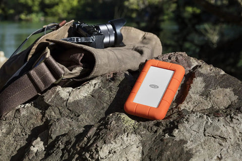 Rugged USB-C Portable External Hard Drive 5TB for Mac and PC, Drop Resistant, placed outdoors on a rock near a camera bag