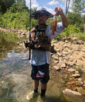 Boy holding a fish with a fishing rod in a stream, wearing a fishing vest and hat, surrounded by rocks and trees.