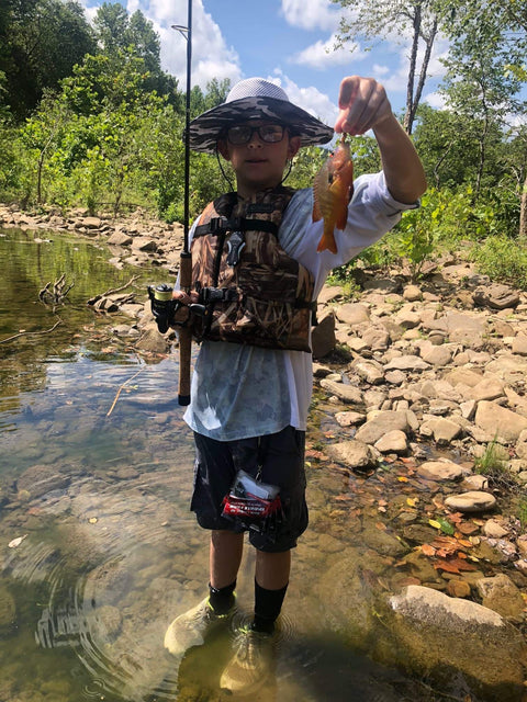 Boy holding a fish with a fishing rod in a stream, wearing a fishing vest and hat, surrounded by rocks and trees.