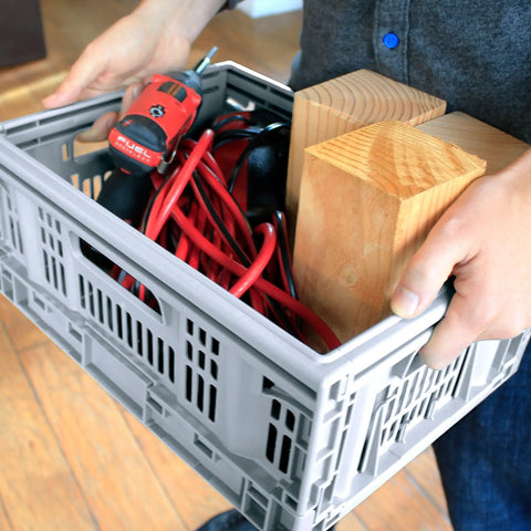 Person carrying a Must-Have Collapsible Utility Crates Set Of 3 Grey 15.8L Stackable Folding Storage Bins filled with tools and wood.