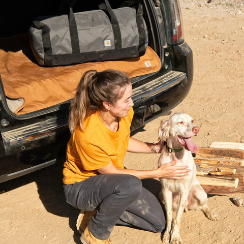 Woman with dog near SUV and Go-To Classic Round Duffel With Utility Pouch, Heavy Duty Water Resistant Packable Gear Bag For Jobsite, Gym, And Travel, 25L Capacity, Rain Defender Protection on the trunk.