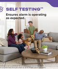 Family sitting on a gray sectional sofa, man using remote control, with text about self-testing smoke and carbon monoxide detectors.