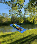 Child enjoying 60 Inch Rectangle Tree Swing With 700 Pound Capacity Open Flat Platform Heavy Duty Waterproof For Kids And Grownups Includes Installation Kit And 2 Tree Straps Blue by a lake.