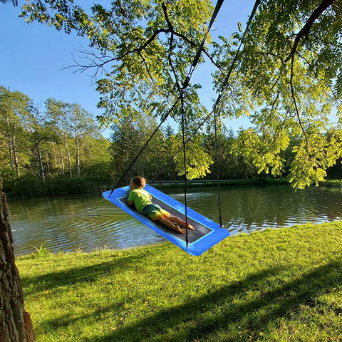 Child enjoying 60 Inch Rectangle Tree Swing With 700 Pound Capacity Open Flat Platform Heavy Duty Waterproof For Kids And Grownups Includes Installation Kit And 2 Tree Straps Blue by a lake.