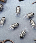 20 Pack Real Deal E10 Miniature Screw Light Bulbs 2.5V 0.3A Tiny Replacement Warm Glow for Toy Cars and Circuit Models Rock Solid E10 Socket Mini Bulb Haul on a grey surface with batteries and tools.