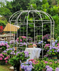 White metal birdcage garden arch arbor, 9 ft high by 6.8 ft wide, surrounded by colorful flowers in an outdoor garden setting.