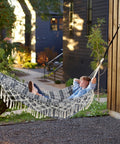 Two Person Indoor Garden Hammock With Tassels And Deluxe Macrame Fringe For Patio Porch Backyard Beach Outdoor And Indoor Cotton Fabric Portable Carry Bag Supports Up To 450 Pounds Blue White, installed outdoors with person relaxing.