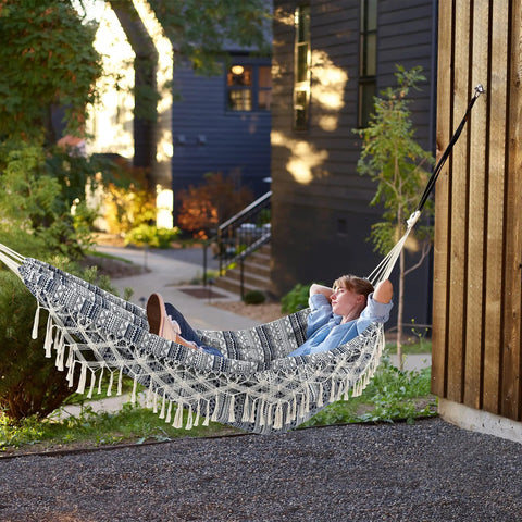 Two Person Indoor Garden Hammock With Tassels And Deluxe Macrame Fringe For Patio Porch Backyard Beach Outdoor And Indoor Cotton Fabric Portable Carry Bag Supports Up To 450 Pounds Blue White, installed outdoors with person relaxing.