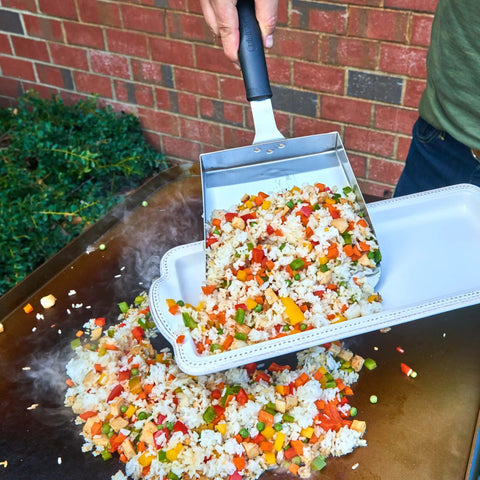 Extra Large Stainless Steel Griddle Food Mover Spatula With 5 Cup Capacity For Rice Vegetables And More Built For Grills And Cookouts Perfect BBQ Tool For Turning Flipping And Transferring A Big Batch Of Food being used to transfer stir-fried rice and vegetables from grill to plate.