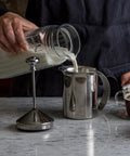Person pouring milk into stainless steel handheld milk frother for creamy foam in seconds, 14 oz capacity.