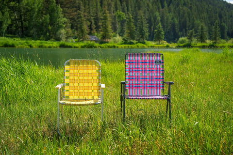Two colorful webbed camping chairs on grass by a lake, one with a yellow pattern and the other a retro webbed camping chair with foldable steel frame.
