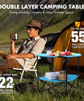 Folding Camping Table Lightweight Double Layer Round Table With 4 Cup Holders And Carry Bag Portable Beach Table For Camping Travel Fishing And Outdoor Activities Blue displayed in use at a campsite near a yellow tent with a person seated holding a cup.