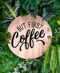 Wooden sign with 'But First Coffee' text surrounded by lush green leaves, emphasizing natural decor.