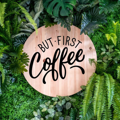 Wooden sign with 'But First Coffee' text surrounded by lush green leaves, emphasizing natural decor.
