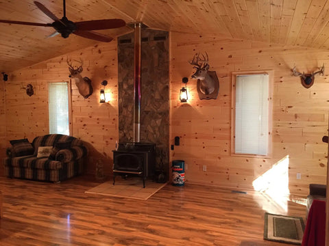 Rustic Wall Sconce Lantern With Glass Globe For Farmhouse Decor And Old School Vibe LED Five Point Five Watt Candle Light Electric Wall Light Fixture In Rust Finish With Authentic Oil Lantern Reproduction Style And Bottom Lit Glow on log cabin wall with wood stove and animal mounts.
