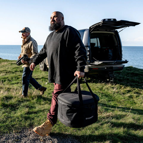 Man holding Venture Bag For Tread Pizza Oven And Roof Rack, Rugged Nylon Carry Case with reinforced top, near parked SUV.