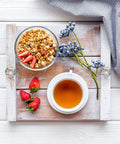 Breakfast tray with granola and strawberries, tea cup, and purple flowers on a wooden tray with diamond knobs on a striped cloth.