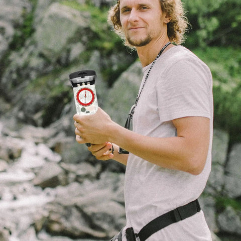 Man holding Orienteering Hiking Compass with ruler and mirror, outdoor navigation tool for camping and survival in natural setting.