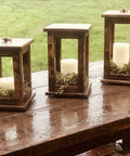Three wooden candle lanterns on a wet table outdoors, showcasing a rustic design and white candles inside.