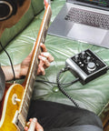 Person using iD4 MkII Audio Interface with Class A Mic Preamps connected to laptop, playing guitar on green sofa.