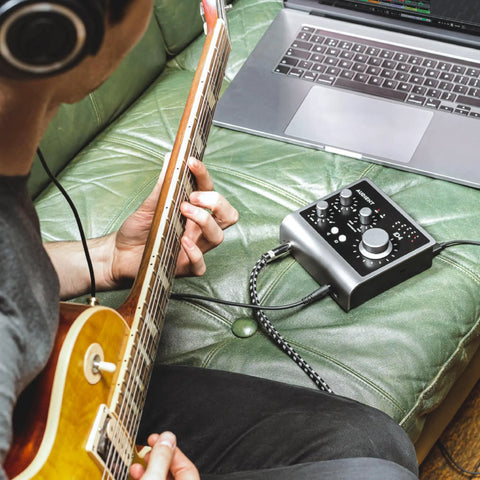 Person using iD4 MkII Audio Interface with Class A Mic Preamps connected to laptop, playing guitar on green sofa.