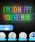 Colorful neon sign with text 'I'm So Happy You're Here' in pink, green, blue, and yellow letters on a brick wall background, USB power options shown.