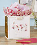 Medium Pink Floral Birthday Gift Bag With Tissue Paper Gold Foil Border Happy Birthday Lettering Laser Cut Flower Accents Satin Ribbon Handles Includes Two Sheets Of Tissue Paper on desk with pen and envelope