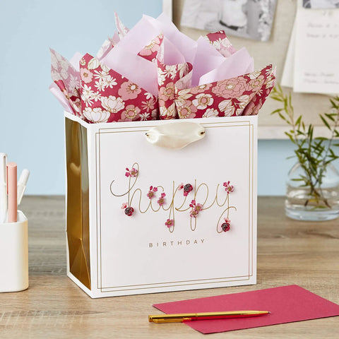 Medium Pink Floral Birthday Gift Bag With Tissue Paper Gold Foil Border Happy Birthday Lettering Laser Cut Flower Accents Satin Ribbon Handles Includes Two Sheets Of Tissue Paper on desk with pen and envelope