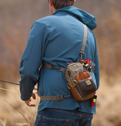 Person wearing Vertical Fly Fishing Chest Pack With Zippered Main Compartment And Interior Pocket Low Profile Tidal Blue Accessory Loops And Belt Loop Attachment For Quick Hands Free Fly Box Storage On The Water, engaged in fishing outdoors.
