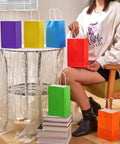 Rainbow Gift Bags With Handles Set 24 Pack 8 Inch Multi Color Paper Party Favor Bags displayed on table with person holding red bag