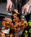 Person using Professional 22 Piece Griddle And BBQ Tool Kit Stainless Steel Dishwasher Safe spatulas to flip shrimp on grill