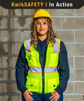 Woman wearing yellow Womens Hi Viz Safety Vest With 9 Pockets, Class 2 Reflective ANSI OSHA Rated, hard hat, standing against wall.