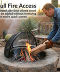 Man placing wood in fire pit with Round Fire Pit Spark Screen Cover With Easy Opening Latch Heavy Duty Steel Mesh Shield For 36 Inch Outdoor Fire Pits Ember Guard.