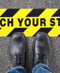 Two Pack Watch Your Step Floor Stickers Anti Slip Safety Decal 6X24 Inch on pavement with black shoes and jeans.