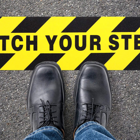 Two Pack Watch Your Step Floor Stickers Anti Slip Safety Decal 6X24 Inch on pavement with black shoes and jeans.