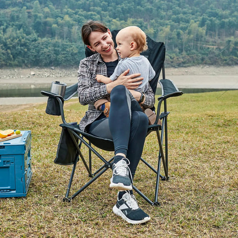 Person sitting in an Oversized Folding Camping Chair With High Back Padded Comfort, outdoors with scenic background, Black.